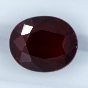 Könnte beinhalten: Ein facettierter, oval geformter Edelstein in tiefroter Farbe. Die Oberfläche des Edelsteins reflektiert das Licht und zeigt seine vielen Facetten und seine innere Struktur. Der Stein scheint ein Rubin zu sein, ein Edelstein, der oft in Schmuck verwendet wird.