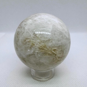 May include: A clear quartz crystal sphere with internal golden inclusions, resting on a clear acrylic stand. The sphere has a polished, reflective surface, showcasing the natural patterns within the stone. This decorative crystal is ideal for home decor.