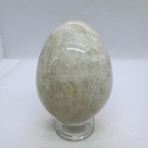 May include: A light-colored, egg-shaped stone sculpture. The stone has a marbled appearance with variations of beige and off-white. The sculpture is displayed on a clear, circular stand, and is set against a white background.