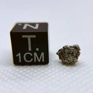 May include: A small, irregularly shaped piece of metallic silver-colored osmium, a rare and dense element, is shown next to a 1 cm cube for scale.