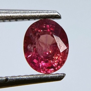 Puede incluir: Una piedra preciosa redonda y roja con un corte facetado. La piedra preciosa se sostiene con unas pinzas.