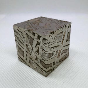 May include: A cube-shaped object with a distinctive patterned surface. The cube is metallic grey with a complex design of intersecting lines and shapes, creating a striking visual texture. The object is likely a decorative piece or a collectible.