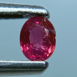 以下が含まれることがあります： 金属製のピンセットで保持された、ファセットのある濃い赤色の楕円形の宝石