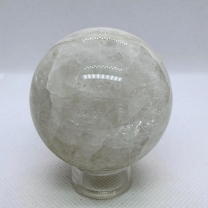 May include: A clear quartz crystal sphere, resting on a clear stand. The sphere has a cloudy, textured appearance with internal veining. The crystal ball is a light, translucent color, reflecting light.