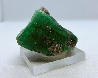 Può includere: Un grande cristallo di smeraldo verde grezzo con alcune inclusioni marroni. Il cristallo è posto su un supporto quadrato trasparente.