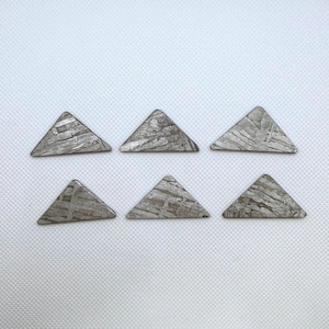 May include: Six triangular, silver-colored meteorite slices. Each slice has a unique, textured pattern with visible lines and variations in tone. The triangles are arranged in two rows of three against a white background.