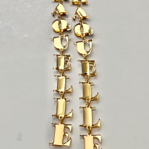 May include: Gold-colored dangle earrings spelling out the word "EQUAL LOVE". The earrings are made of a shiny, reflective metal and feature a series of connected letters. The letters are in a bold, sans-serif font.
