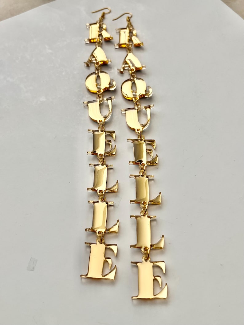 May include: Gold-colored dangle earrings spelling out the word "EQUAL LOVE". The earrings are made of a shiny, reflective metal and feature a series of connected letters. The letters are in a bold, sans-serif font.