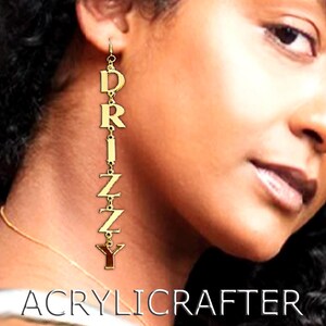 May include: Gold-colored dangle earrings spelling out "DRIZZY" in capital letters. The earrings are suspended from a hook and are connected by small bars. The word "ACRYLICRAFTER" is printed in white at the bottom of the image.