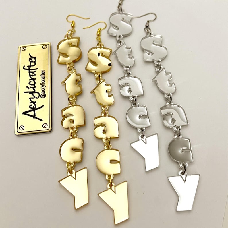 May include: Gold and silver-toned dangle earrings spelling out "STACY" in a bold, blocky font. The earrings have a hook closure and are displayed with a gold metal tag that reads "Acrylicrafter" and "@acrylicrafter".