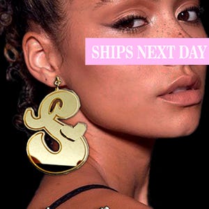 May include: Gold-tone oversized letter L earrings. The earrings are in the shape of a stylized letter L. The earrings are shown on a model wearing a black top.