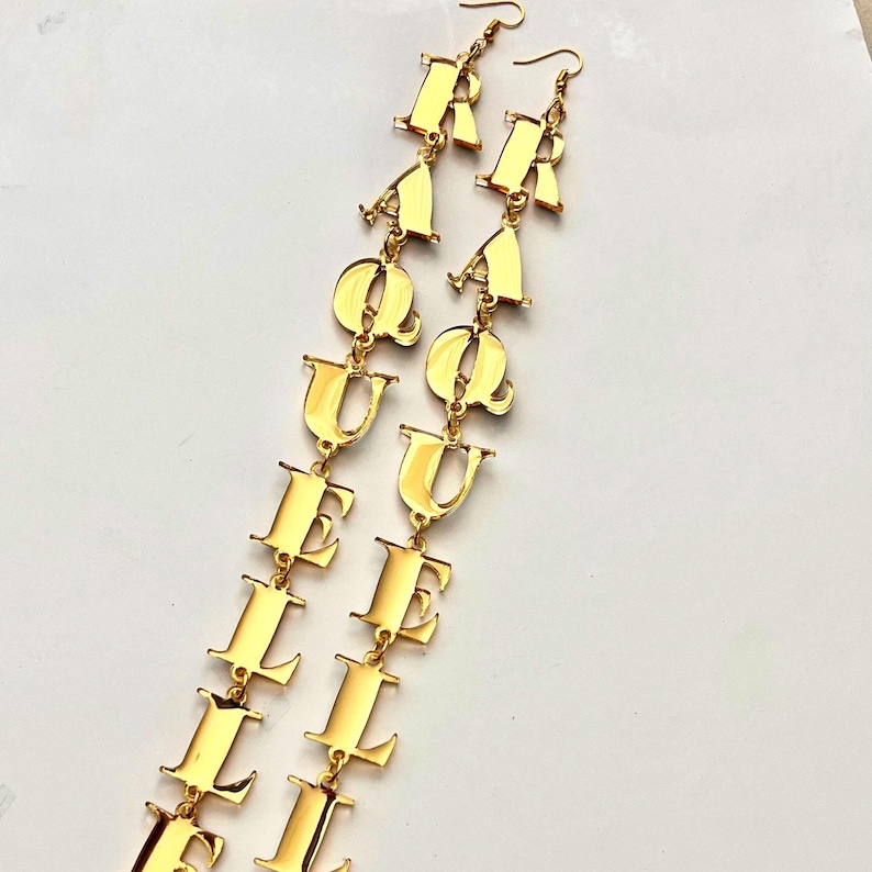 May include: A pair of gold-colored dangle earrings spelling out the word "RAQUEL" in capital letters. The earrings are made of a shiny, reflective material and hang from simple ear hooks. The letters are connected by small links.
