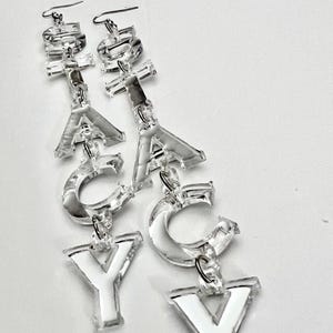 May include: Clear acrylic earrings spelling out the word "YAY". Each earring features a fish hook and is composed of individual letters. The letters are clear with white accents, creating a bold and playful accessory.