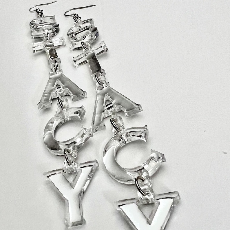 May include: Clear acrylic earrings spelling out the word "YAY". Each earring features a fish hook and is composed of individual letters. The letters are clear with white accents, creating a bold and playful accessory.
