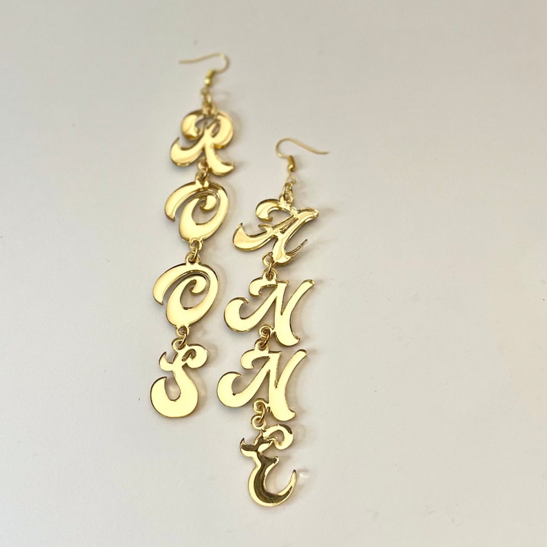 May include: Gold-colored statement earrings spelling out the word "ROSEANNE" in a cursive font. The earrings are made of a shiny, reflective material and hang from a simple hook. The letters are connected, creating a dangling effect.