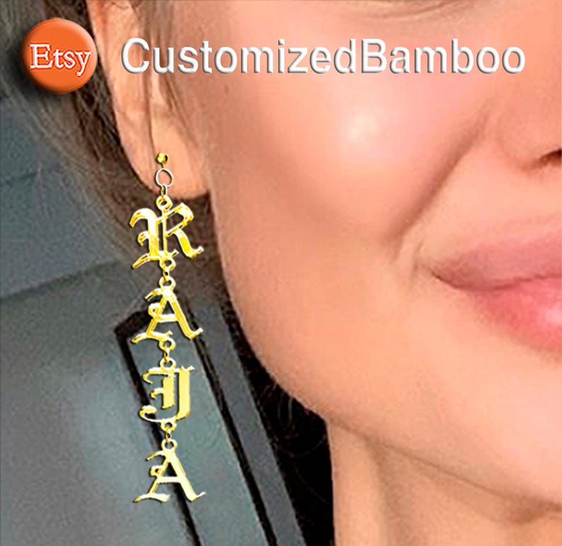 May include: Gold-colored dangle earrings spelling out the name "RAJA" in an elegant, old English font. The earrings are suspended from a small hoop and are shown against a blurred background. The Etsy logo and "CustomizedBamboo" text are in the upper left.