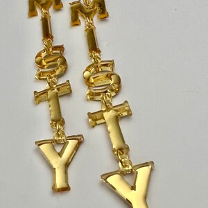 May include: A pair of gold-colored statement earrings spelling out the letters "M", "S", "T", and "Y". The earrings are made of a shiny, reflective material and hang from gold-colored ear wires. The letters are connected by small gold links.