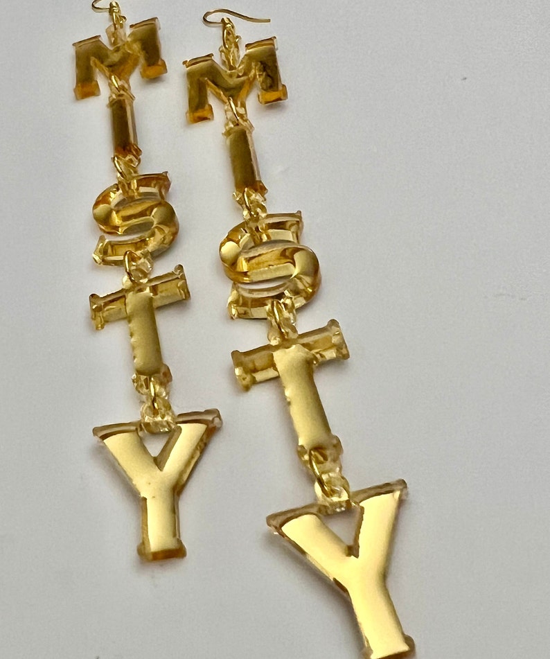 May include: A pair of gold-colored statement earrings spelling out the letters "M", "S", "T", and "Y". The earrings are made of a shiny, reflective material and hang from gold-colored ear wires. The letters are connected by small gold links.