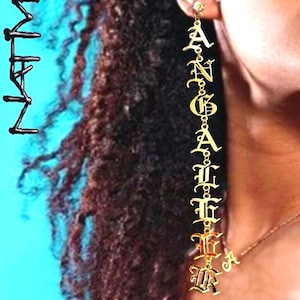 May include: Gold-colored dangle earrings spelling out the name "ANGELICA" in a gothic font. The earrings are long and hang vertically. The background shows a person's hair and skin.