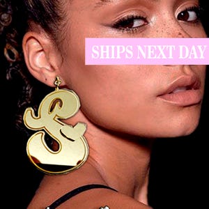 May include: Gold-colored statement earrings in the shape of the letter "&" with a heart detail. The earrings are large and reflective, with a shiny finish. The image includes the text "SHIPS NEXT DAY".
