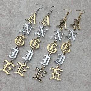 May include: Four pairs of dangle earrings spelling out the word "GANG" in a gothic font. The earrings alternate between gold and silver tones, with the letters connected by small chains and hooks.