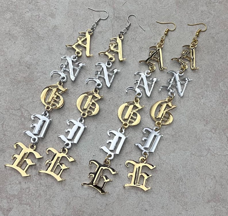 May include: Four pairs of dangle earrings spelling out the word "GANG" in a gothic font. The earrings alternate between gold and silver tones, with the letters connected by small chains and hooks.