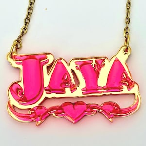 May include: A gold-toned metal nameplate necklace featuring the name JAYA in bold, stylized lettering. The letters are filled with a vibrant, translucent hot pink acrylic material. A small pink heart is centered below the name.