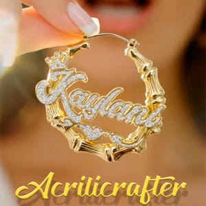 May include: A gold-toned bamboo hoop earring featuring the name Kaylani in cursive script. The name is encrusted with clear stones, and a small crown sits above the first letter. The hoop has a textured bamboo-like design with a metallic finish.