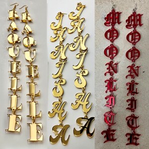 May include: Three pairs of statement earrings. Two pairs are gold-colored, spelling out names. The third pair is red, with a gothic font. All earrings are long and dangle from a hook.