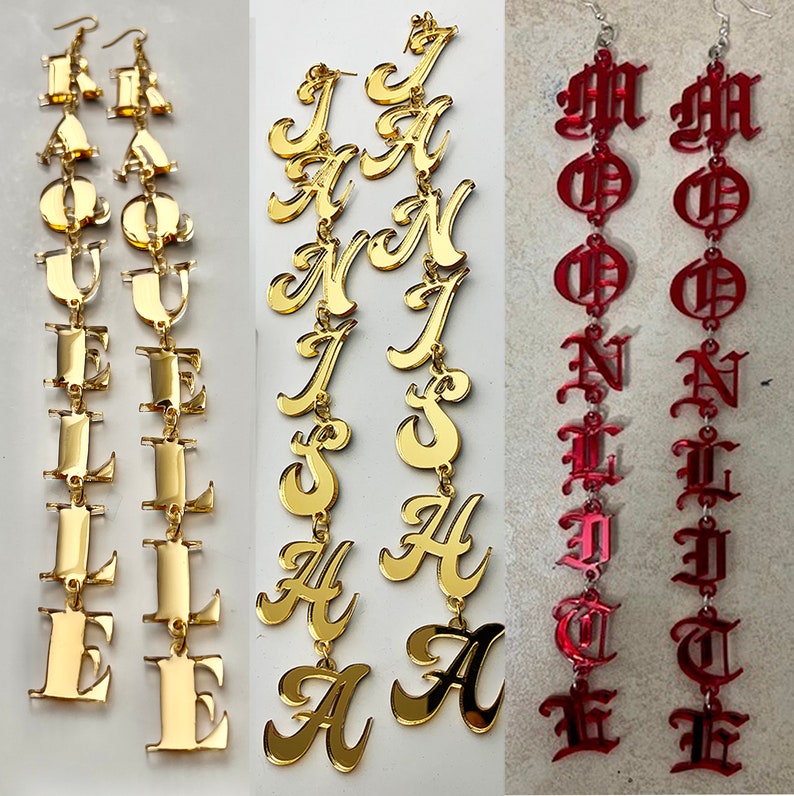 May include: Three pairs of statement earrings. Two pairs are gold-colored, spelling out names. The third pair is red, with a gothic font. All earrings are long and dangle from a hook.