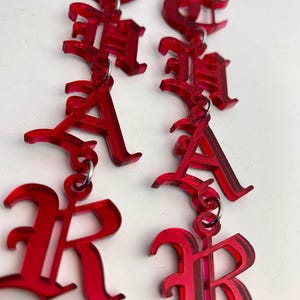 May include: A pair of red acrylic earrings spelling out the word "RACHA" in a gothic font. The earrings are suspended from silver-colored ear wires and feature a layered design for a 3D effect. The letters are connected by small metal rings.