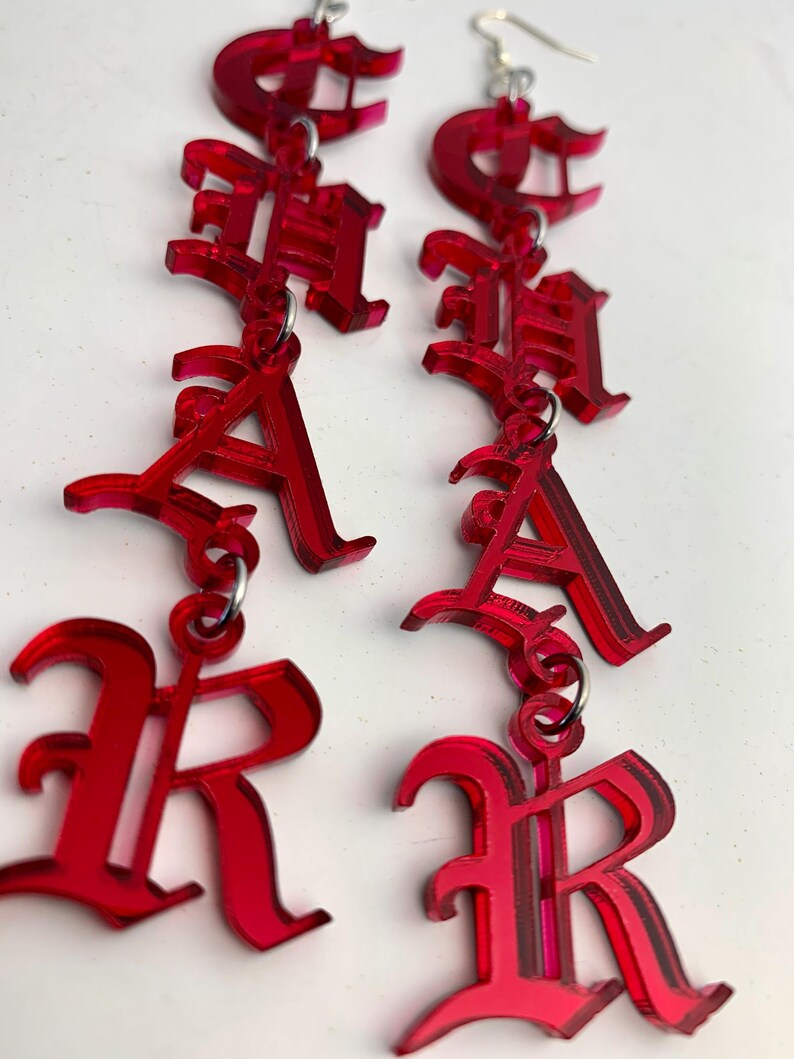 May include: A pair of red acrylic earrings spelling out the word "RACHA" in a gothic font. The earrings are suspended from silver-colored ear wires and feature a layered design for a 3D effect. The letters are connected by small metal rings.