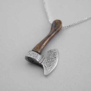 Puede incluir: Un colgante de hacha de plata con un mango de madera marrón en una cadena de plata. La cabeza del hacha tiene una superficie texturizada.