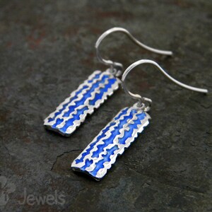 Pode incluir: Um par de brincos de prata com um design de esmalte azul. Os brincos têm um aspeto texturizado, martelado e têm a forma de um retângulo.