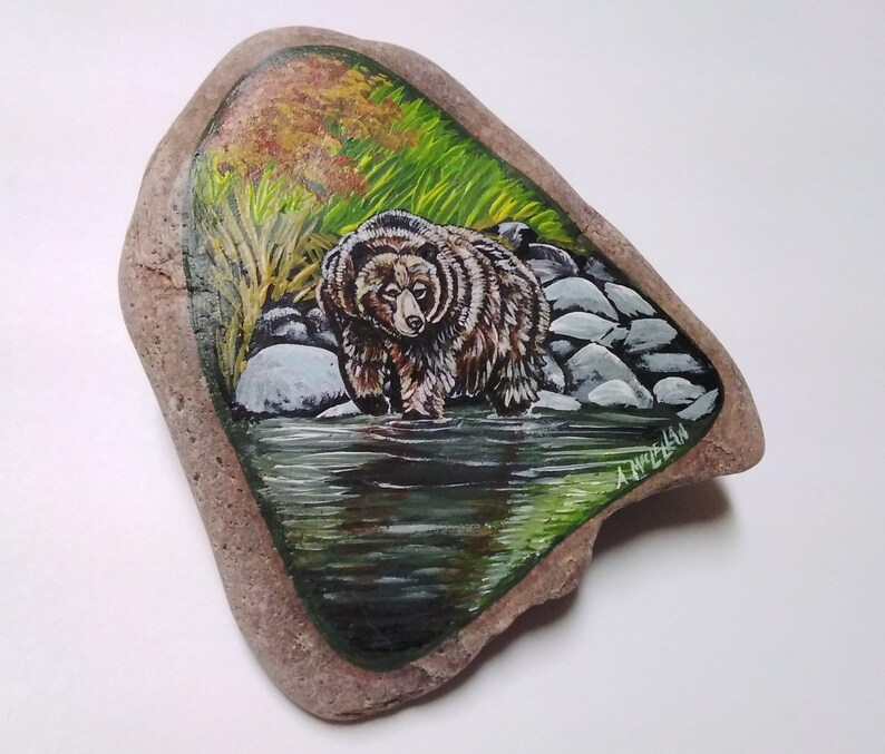 May include: Hand-painted rock art featuring a brown bear standing in a green stream with rocks and foliage. The artist's signature, "A. MELLAN", is visible in the lower right corner.