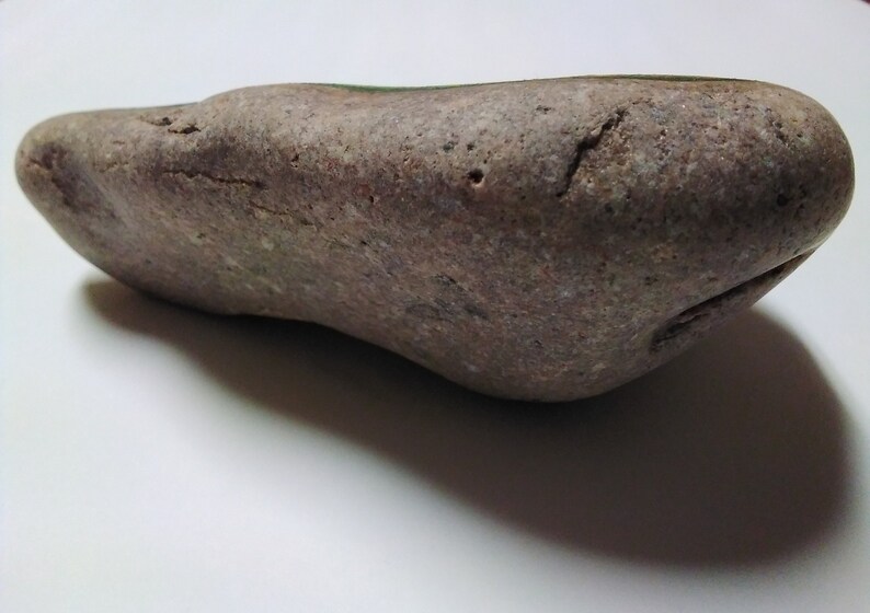May include: A smooth, gray, and brown rock with a slightly green hue. The rock is oval-shaped and has a rough texture.