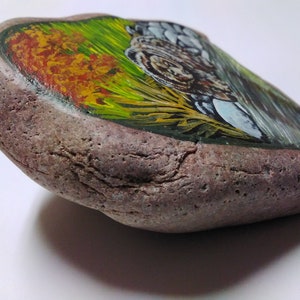 May include: A painted rock with a landscape scene of a forest with green and yellow grass, a brown animal, and a blue sky.