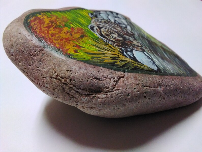 May include: A painted rock with a landscape scene of a forest with green and yellow grass, a brown animal, and a blue sky.