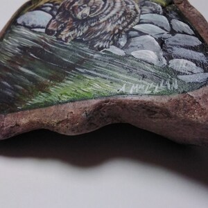May include: Hand-painted rock art featuring a brown bear walking through a green forest with gray rocks. The artist's signature, "A. Mitchell", is visible in the lower right corner.