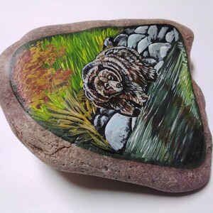 May include: Hand-painted rock art featuring a brown bear peeking out from behind a green leafy bush and rocks. The background is a mix of green, yellow, and orange.