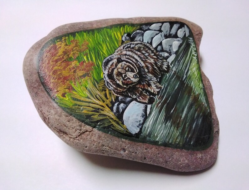 May include: Hand-painted rock art featuring a brown bear peeking out from behind a green leafy bush and rocks. The background is a mix of green, yellow, and orange.