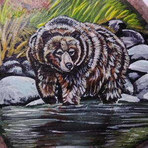 May include: A hand-painted rock art piece depicting a brown bear standing in a river with green grass and rocks in the background.