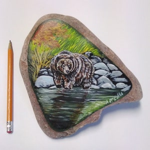 May include: Hand-painted rock art featuring a brown bear standing in a stream with green grass and rocks. The artist's signature is visible in the lower right corner.