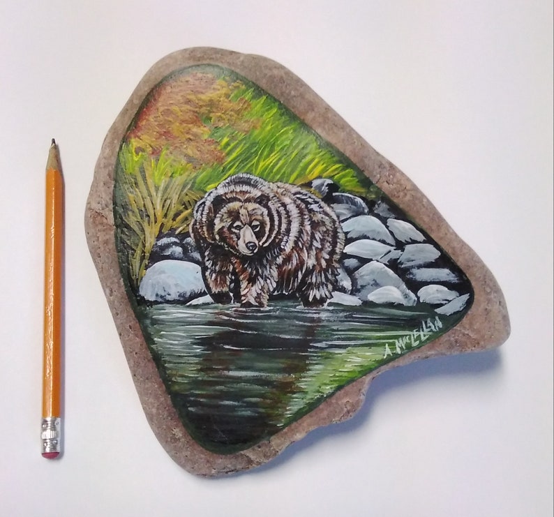 May include: Hand-painted rock art featuring a brown bear standing in a stream with green grass and rocks. The artist's signature is visible in the lower right corner.