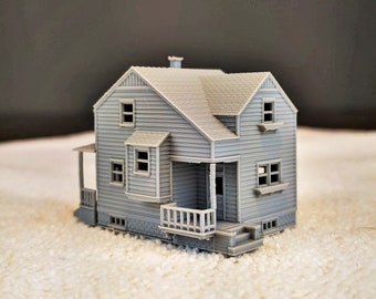 Op de afbeelding: Een grijs 3D-geprint model van een klein huis met een veranda en trappen. Het huis heeft een zadeldak en ramen aan de voor- en zijkanten.