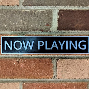May include: A black rectangular sign with silver lettering that reads "NOW PLAYING" mounted on a brick wall.