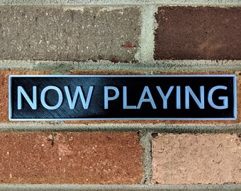 May include: A black rectangular sign with silver lettering that reads "NOW PLAYING" mounted on a brick wall.