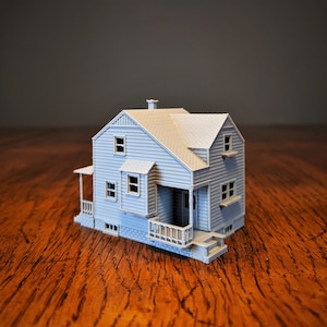 May include: A gray model house with a white porch and a small front yard. The house has a gabled roof and a chimney. The model is made of plastic and is about 6 inches tall.