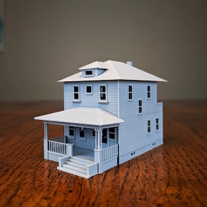 Op de afbeelding: Een lichtblauw modelhuis met een veranda, trappen en een gedetailleerd dak. De twee verdiepingen tellende structuur heeft meerdere ramen en een schoorsteen. Het huis staat op een houten oppervlak.