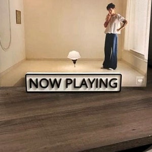 May include: A white sign with black text that reads "NOW PLAYING" on a brown wooden surface.